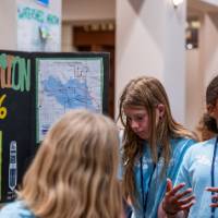 three 6th grade students in light blue T-shirts that say "Living Stones Academy" on them, students talking and discussing their posters to an audience off-camera